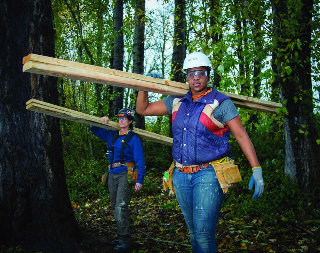 Two women dressed for construction in a forest carrying lumber
