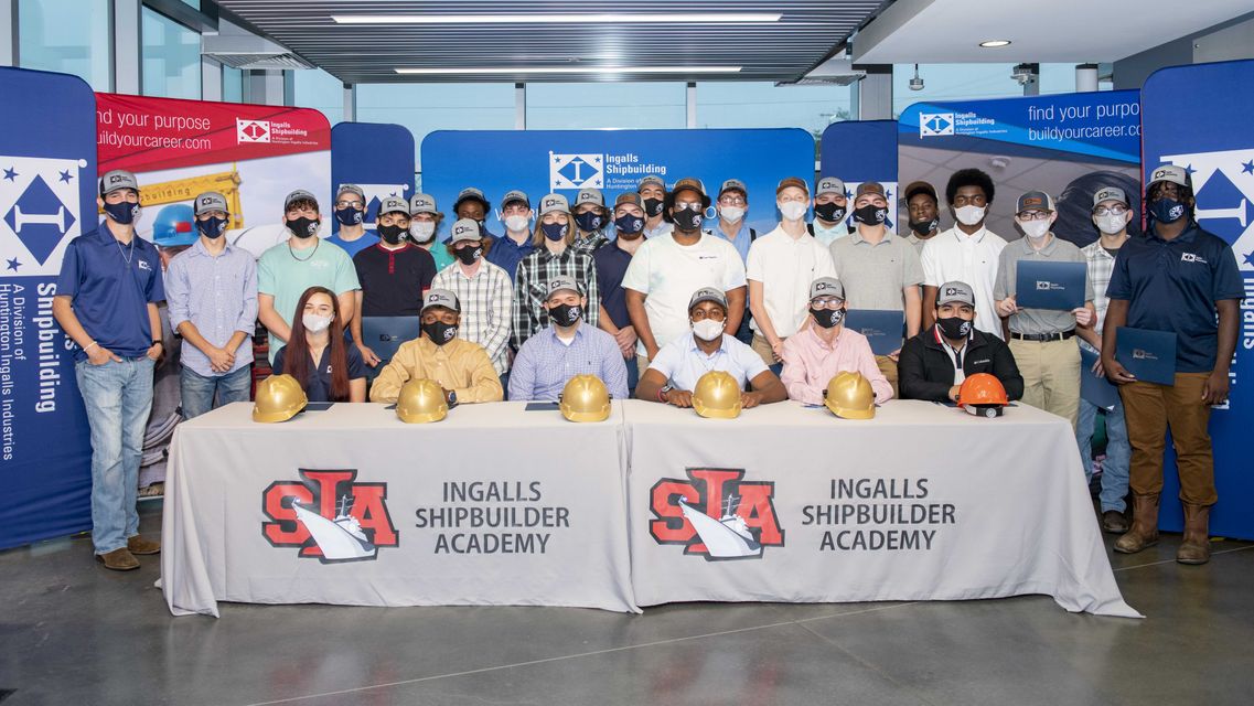 29 high school seniors in masks posing behind table with Ingalls Shipbuilding Academy logo