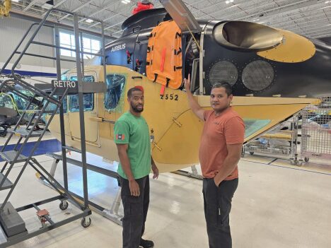 Two men stand proudly beside an Airbus helicopter in a hangar.